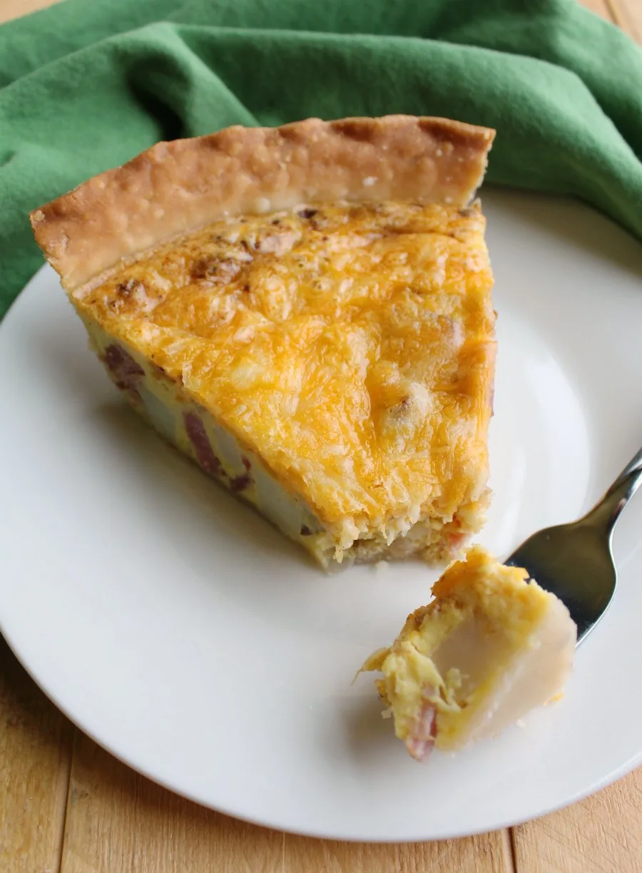 Slice of ham, potato and cheese quiche with first bite on fork.