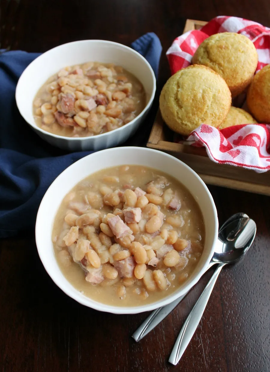 Bowls of thick ham and bean soup next to a couple of spoons and a basket of cornbread muffins.