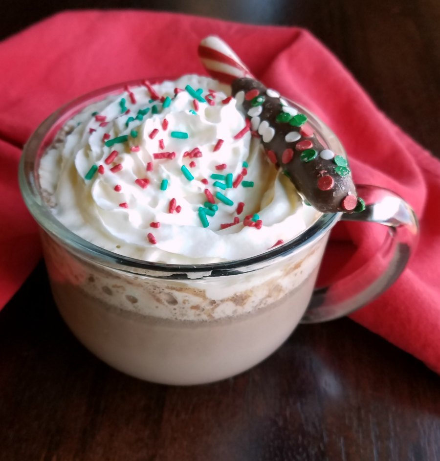 glass mug of creamy hot cocoa topped with whipped cream, sprinkles and a chocolate dipped peppermint stick.