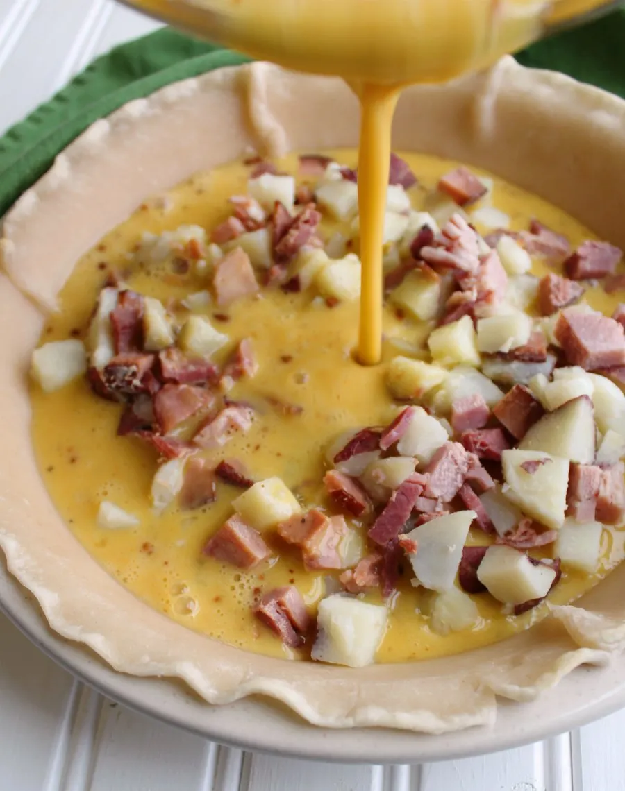 pouring egg mixture into pie shell filled with ham and potatoes.