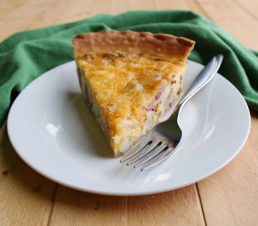 Slice of ham, potato and cheese quiche on plate with fork.