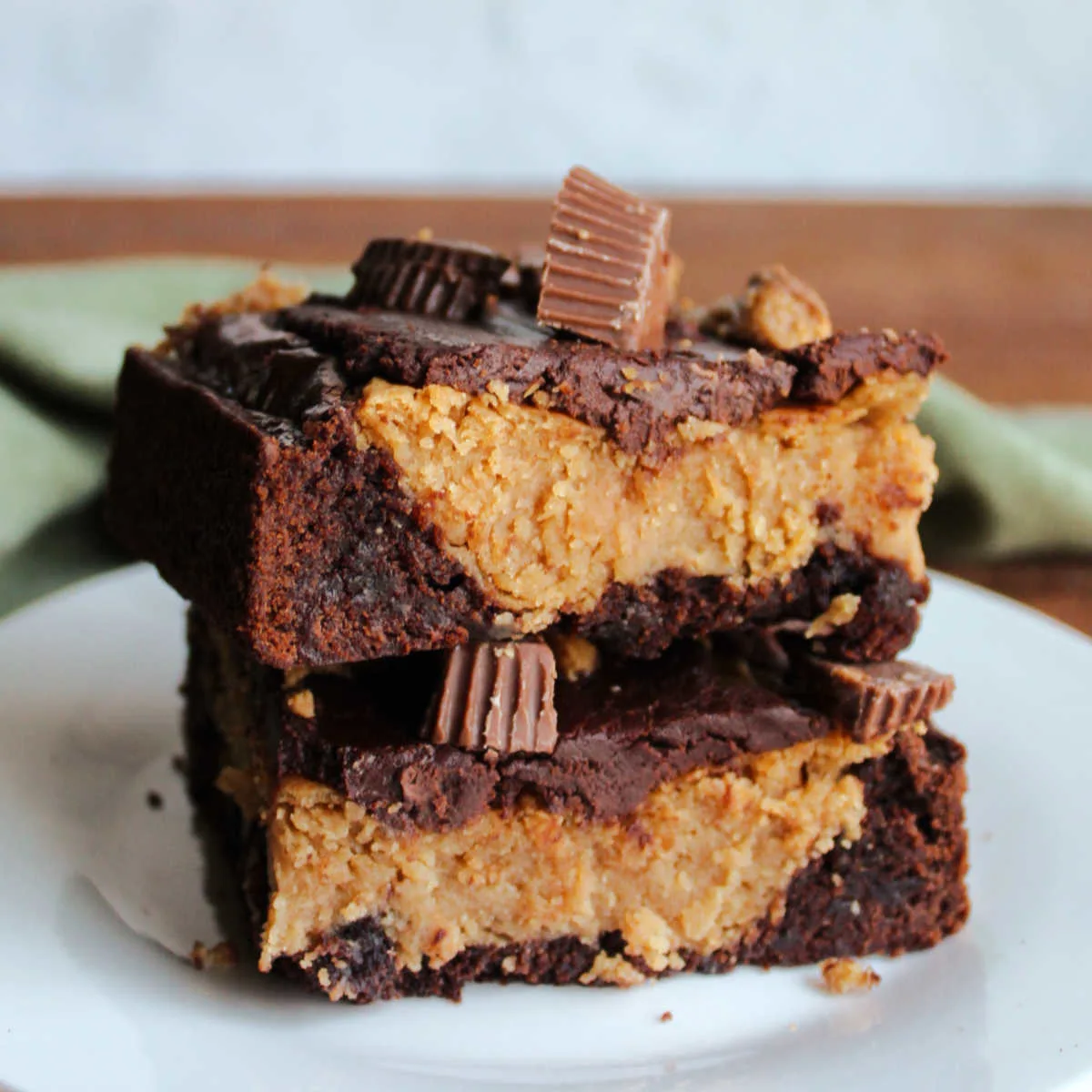 Two slices of peanut butter cheesecake brownies stacked on top of each other, showing rich brownie bottom layer, creamy peanut butter cheesecake layer, topped with chocolate ganache and chopped peanut butter cup candies.