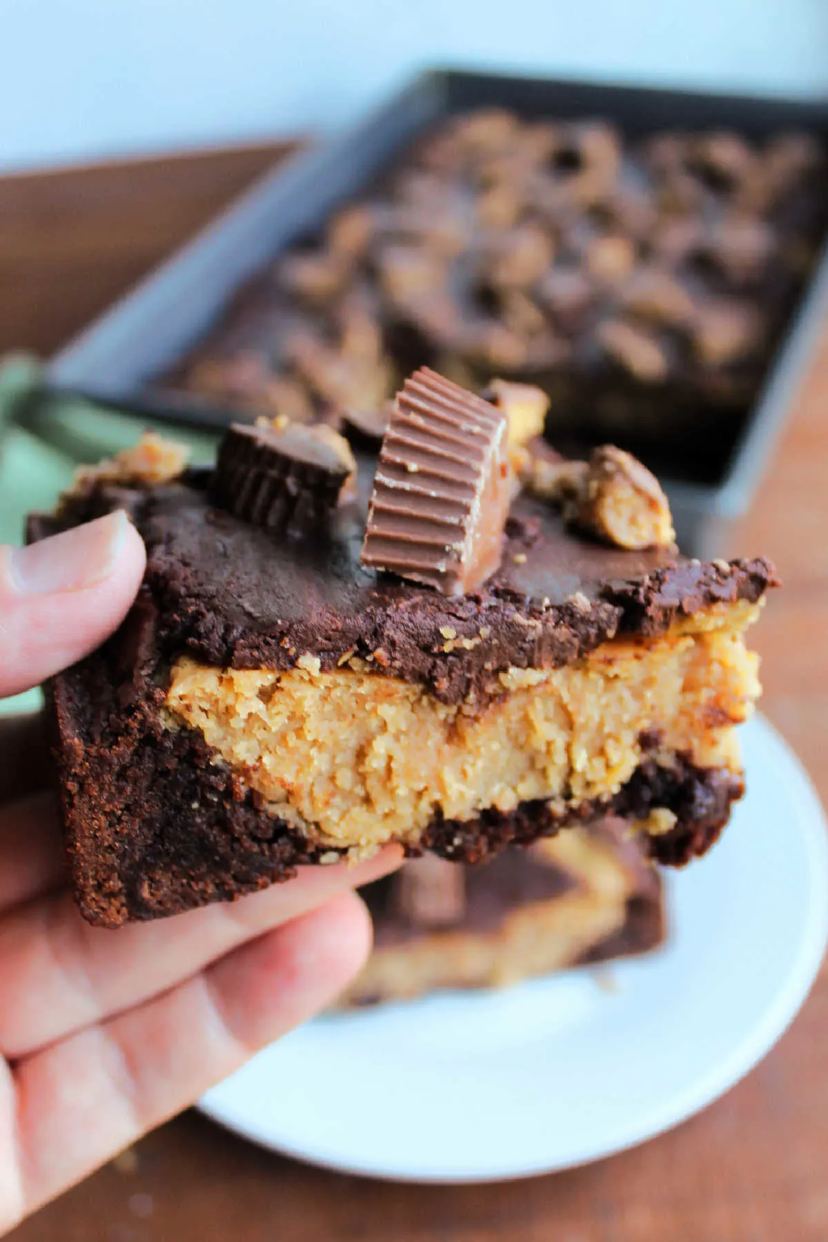 Hand holding piece of brownie with peanut butter cheesecake filling, chocolate topping, and chopped peanut butter cup candies on top, ready to eat. 