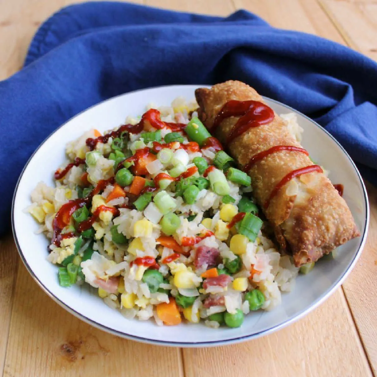Bowl of pork fried rice topped with sriracha and an egg roll.