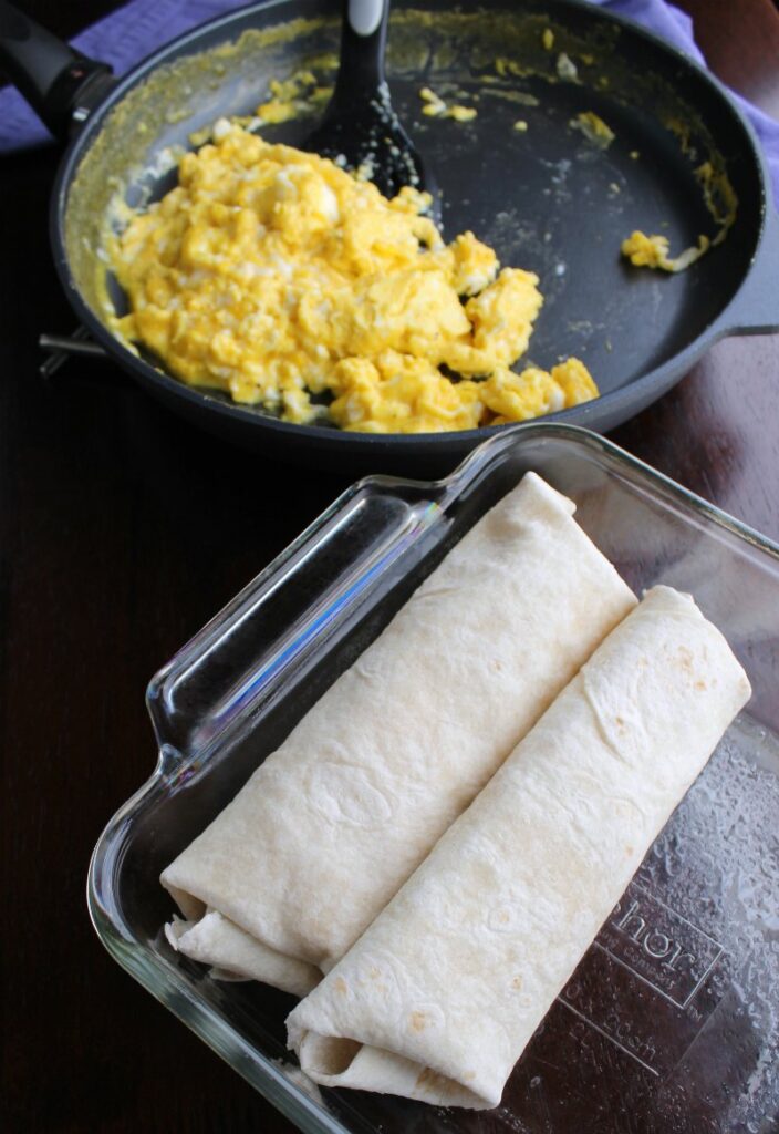 rolled burritos in pan in front of pan of remaining scrambled eggs.