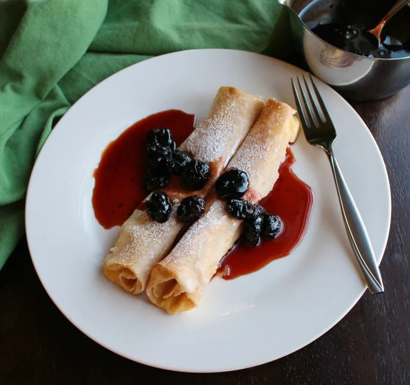 Two baked lemon ricotta stuffed crepes topped with powdered sugar and cherry sauce.