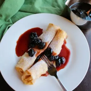 fork cutting into stuffed crepe covered with cherry syrup. pan of syrup in background.