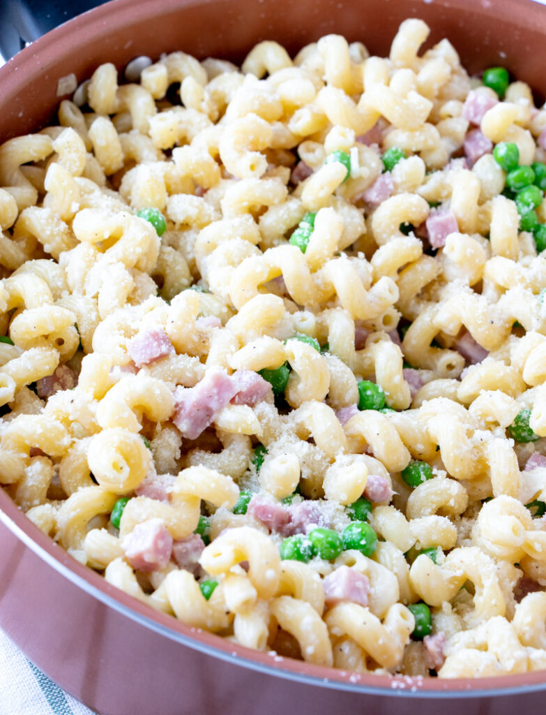 Sauté pan filled with curly pasta, chunks of ham and green peas in a light cream sauce.