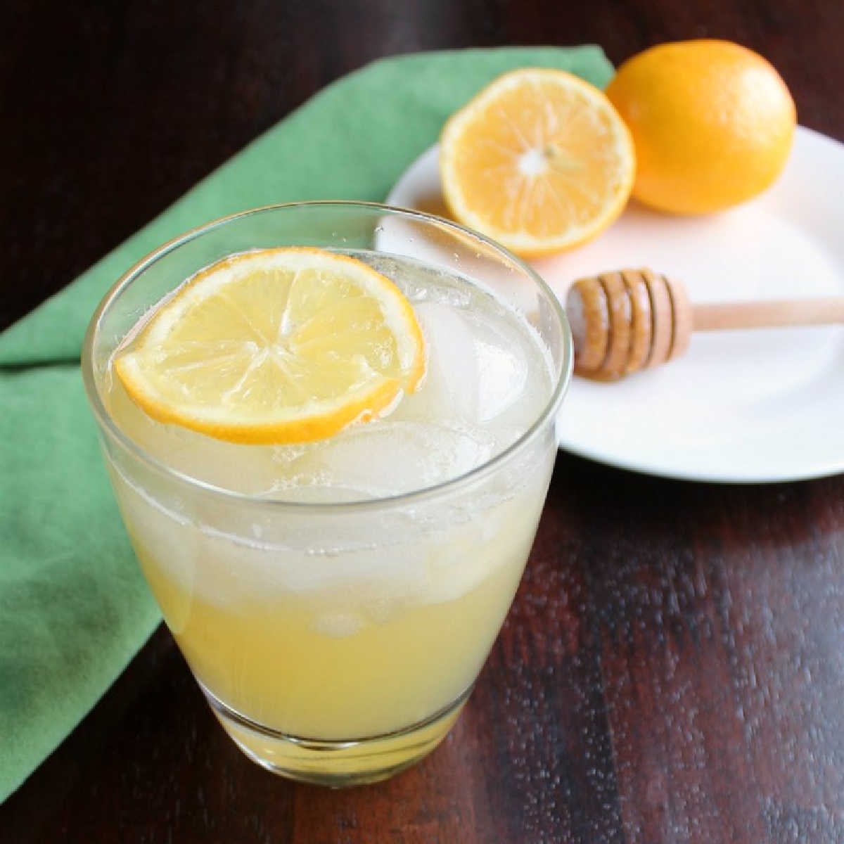 glass of honey lemonade on ice with floating lemon slice with honey drizzler and additional fresh lemons in background.