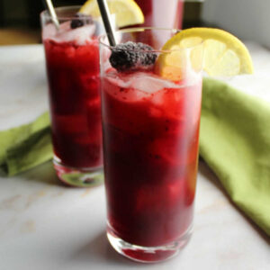 Glasses of dark purple lemonade with lots of pureed blackberries and ice inside topped with frozen blackberries and a slice of lemon.