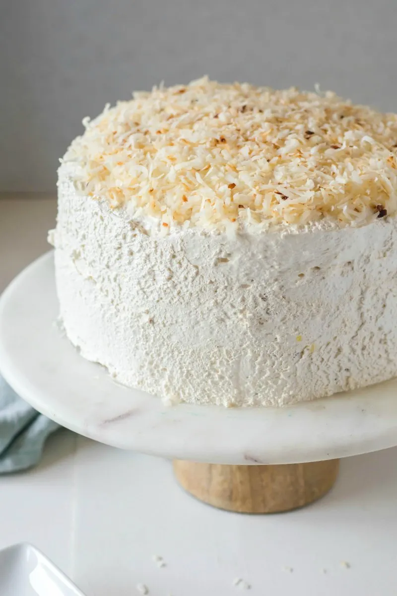 Round layer cake with fluffy white frosting and toasted coconut on top on cake stand.