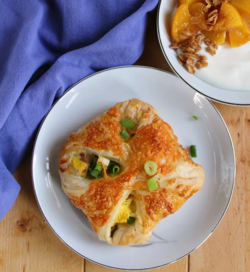 ham egg and cheese hand pie with scallions on top and a bowl of yogurt with peaches and granola in the background.