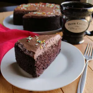 slice of chocolate cake with remaining cake and coffee cup in the background.