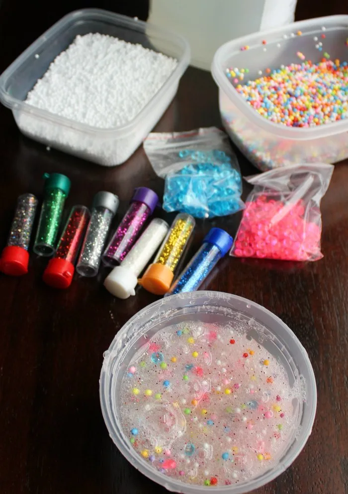 tub of slime next to tubes of glitter, plastic beads, foam balls and gallon of clear glue.