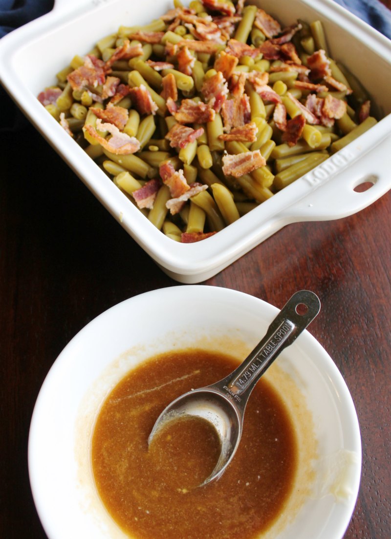 pan of green beans with bacon on top in background with bowl of sauce mixture for smothered green beans, ready to smother and cook