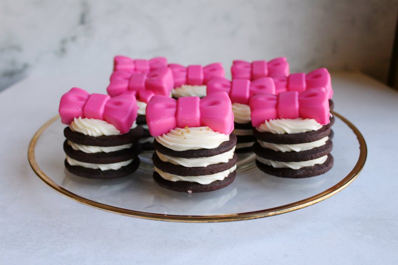 Plate of chocolate sandwich cookie stacks with layers of dark chocolate cookies with white frosting between topped with pink chocolate bows.