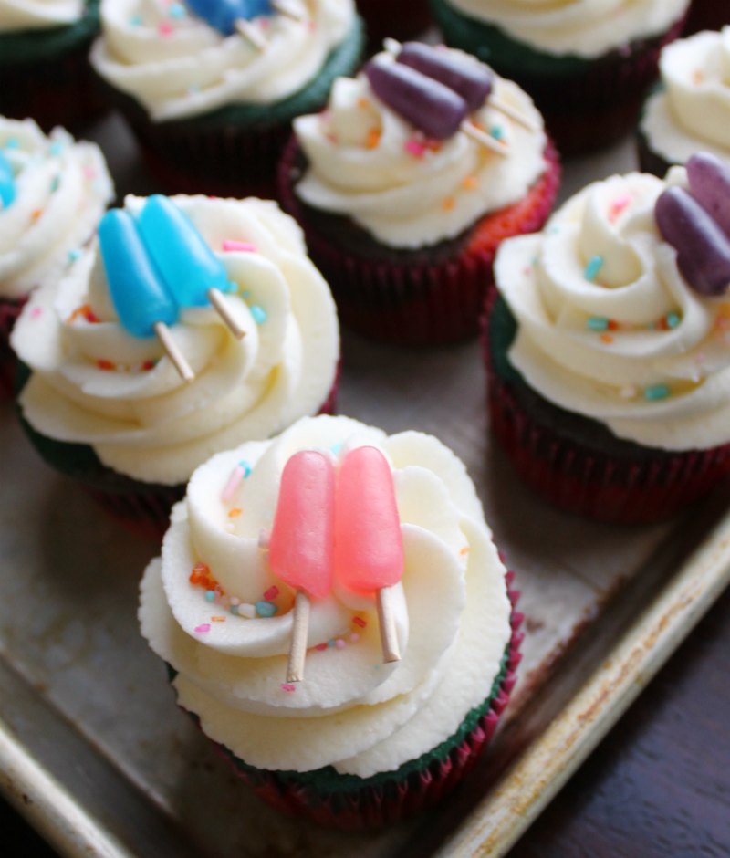 tray of cupcakes with swirls of cream cheese frosting, sprinkles and candy cupcake toppers that look like popsicles