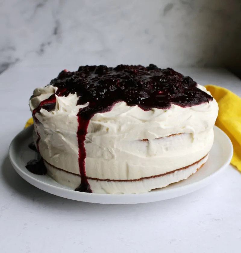 whole blackberry cake with lots of fluffy white frosting on top, barely frosted sides and some blackberry sauce dripping over the edge.