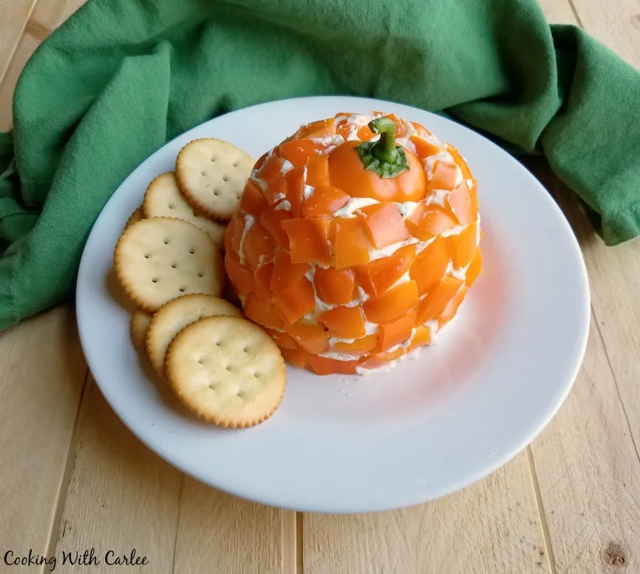 Havarti cheese ball coated in orange bell pepper pieces with the top of a bell pepper on top as a pumpkin stem with crackers on a plate.