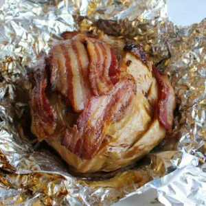 whole head of cabbage wrapped in bacon fresh off the grill and partially wrapped in foil.