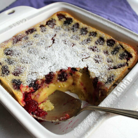 Blackberry Clafoutis 3 square pan on blackberry clafoutis with berry spoon in corner and golden custardy inside showing.