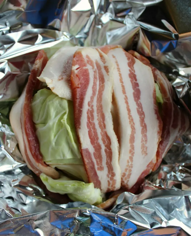 head of cabbage wrapped in bacon slices with aluminum foil around it