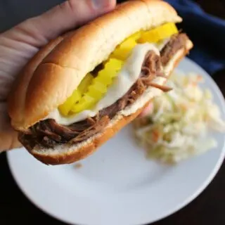 Hand holding Italian beef sandwich with shredded beef, melted provolone cheese, and pepperoncini slices, ready to eat.