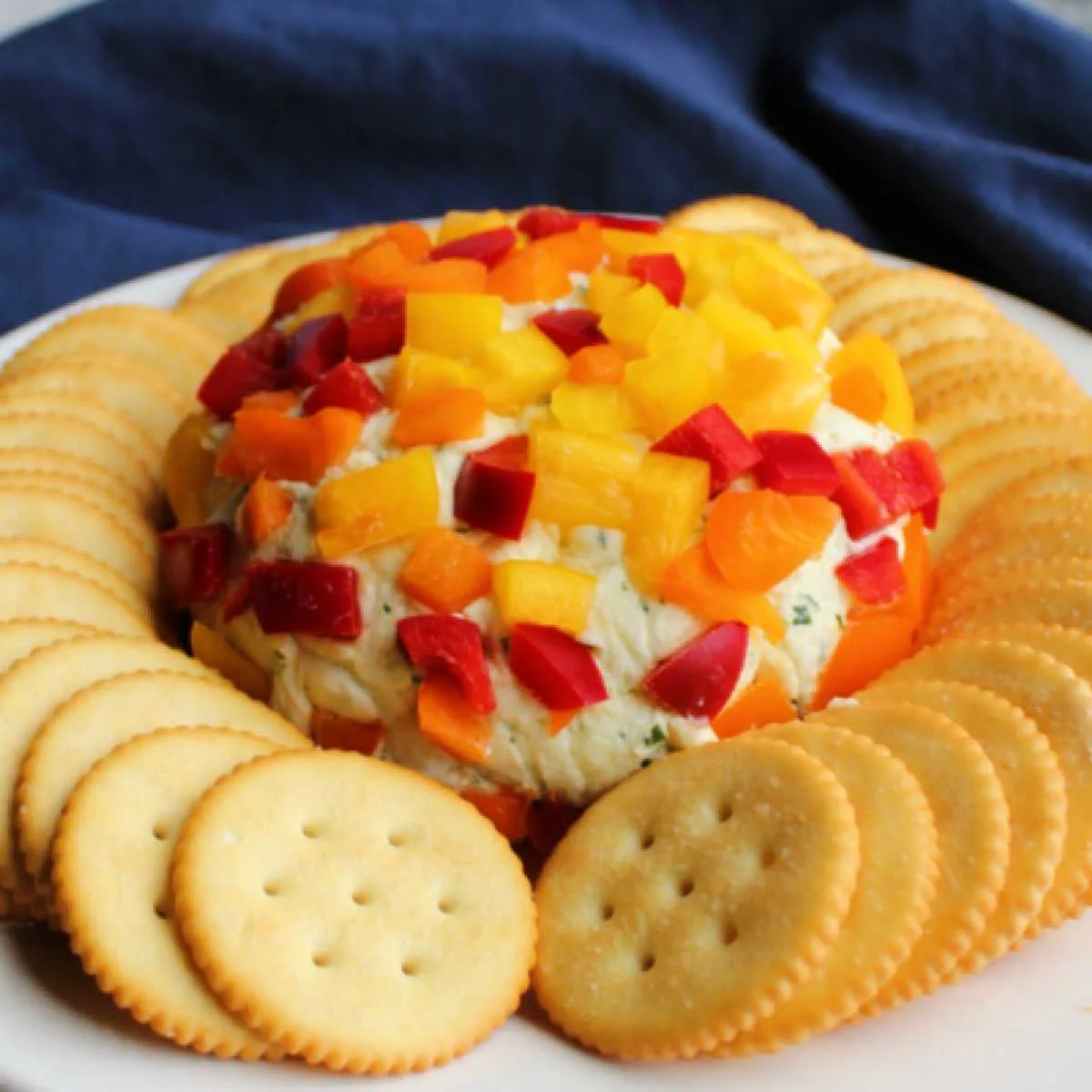 Creamy havarti cheese ball coated in diced red, yellow, and orange bell pepper on plate surrounded by crackers, ready to be served.