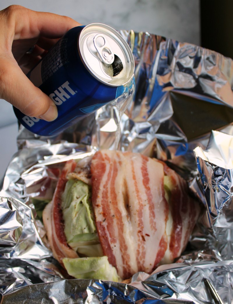 pouring beer over bacon wrapped head of cabbage in foil bowl.