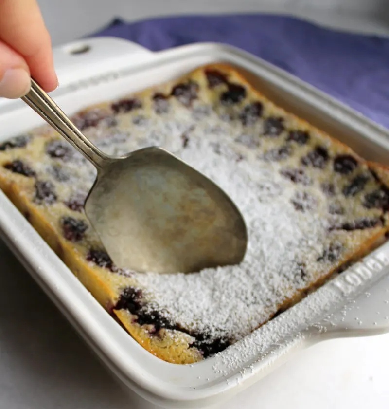 berry spoon going into pan of blackberry clafoutis covered with powdered sugar.