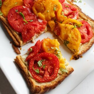 Square piece of tomato tart with golden brown flaky puff pastry crust topped with ricotta cheese layer, red and yellow tomato slices and bits of fresh basil.