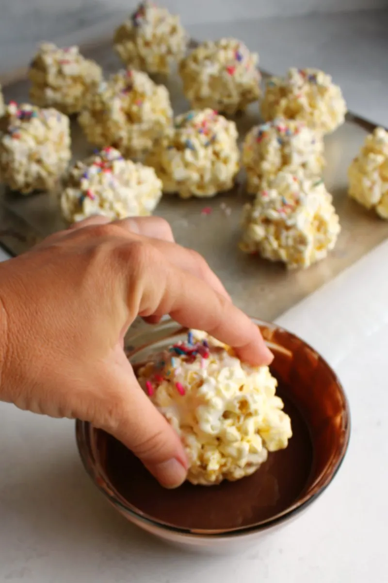 hand dunking marshmallow popcorn ball into bowl of melted milk chocolate.