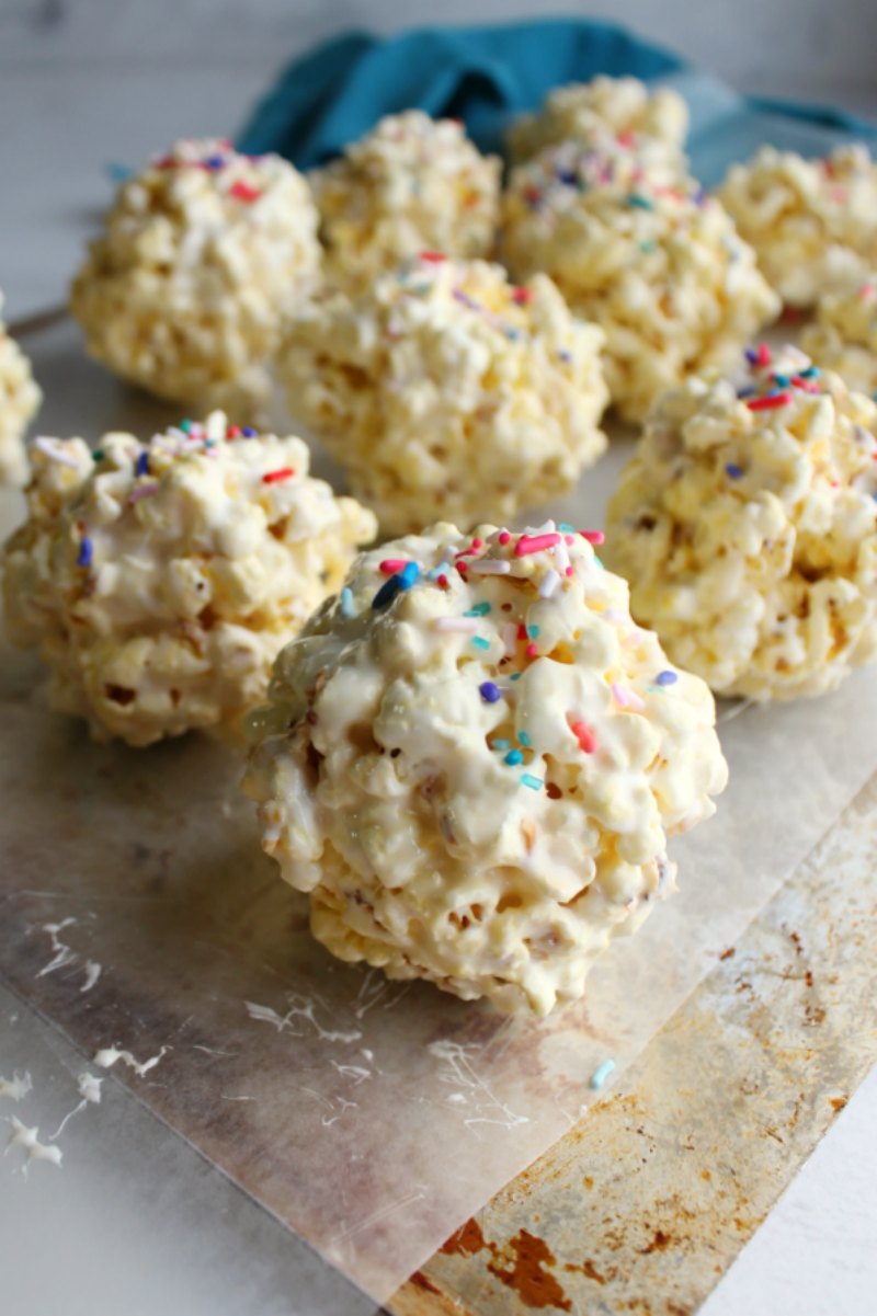 close up of marshmallow popcorn balls with sprinkles on top sitting on wax paper lined baking tray.