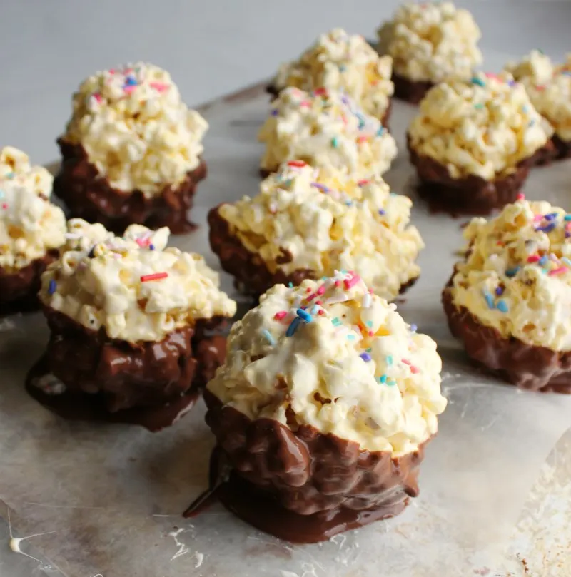 Marshmallow popcorn balls with the bottom halves dipped in chocolate and sprinkles on top setting up on a sheet of wax paper.