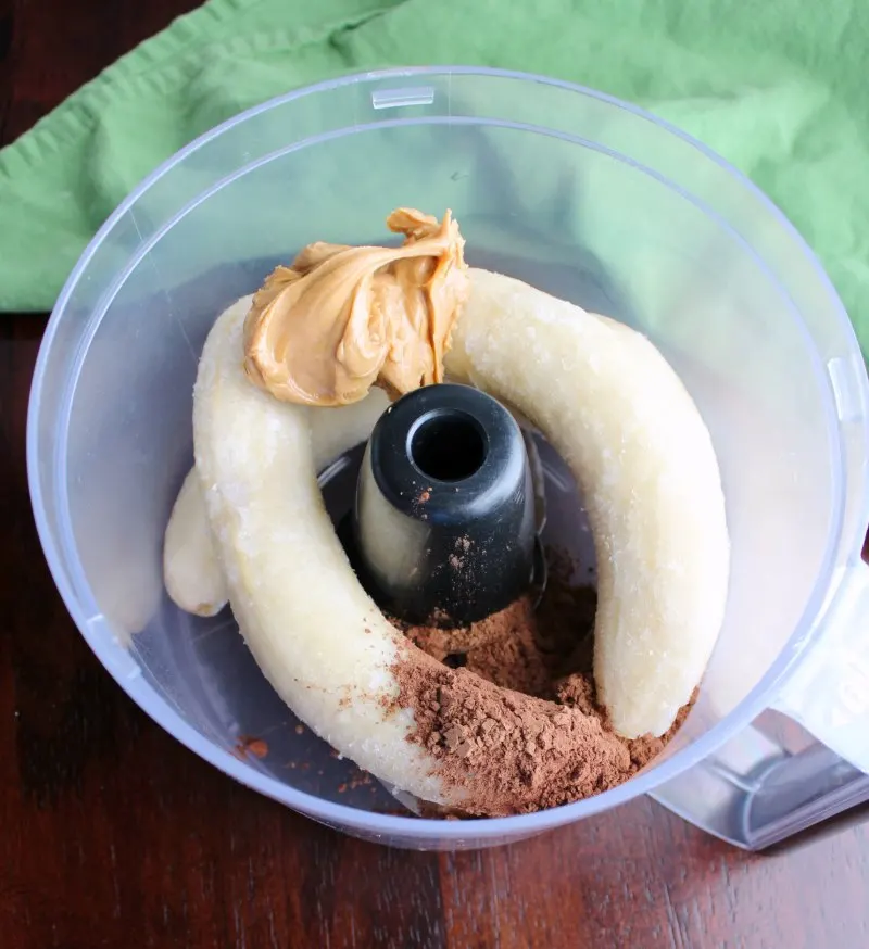 food processor bowl with frozen bananas, peanut butter and cocoa powder for nice cream.