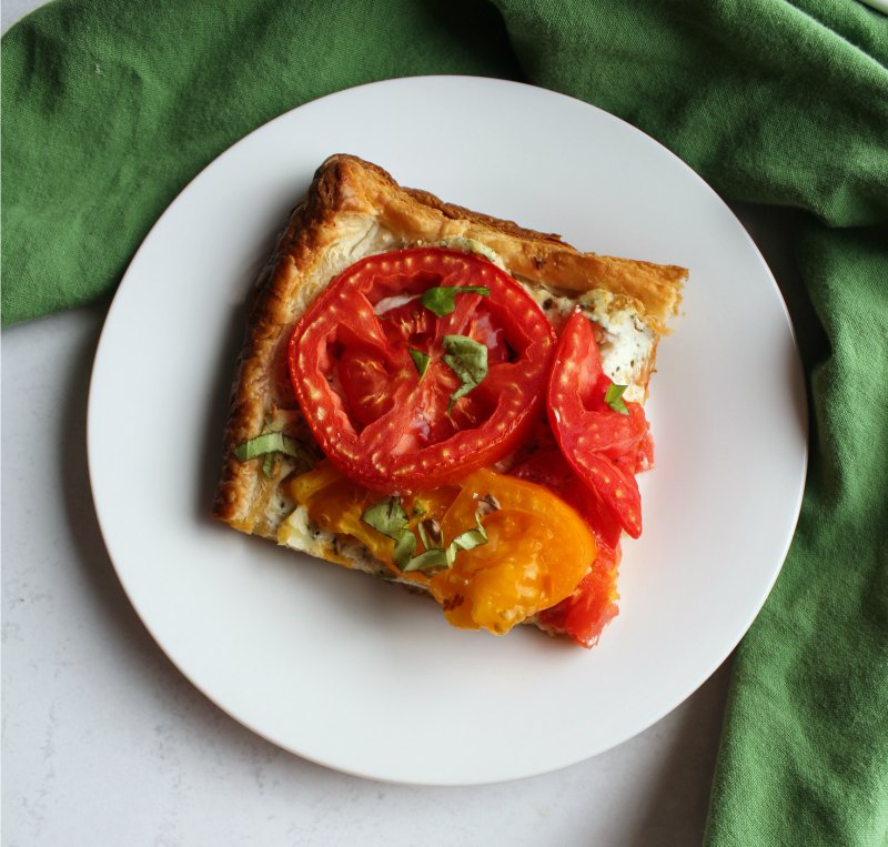slice of tomato tart with red and yellow tomatoes, puff pastry, ricotta and basil served on plate..