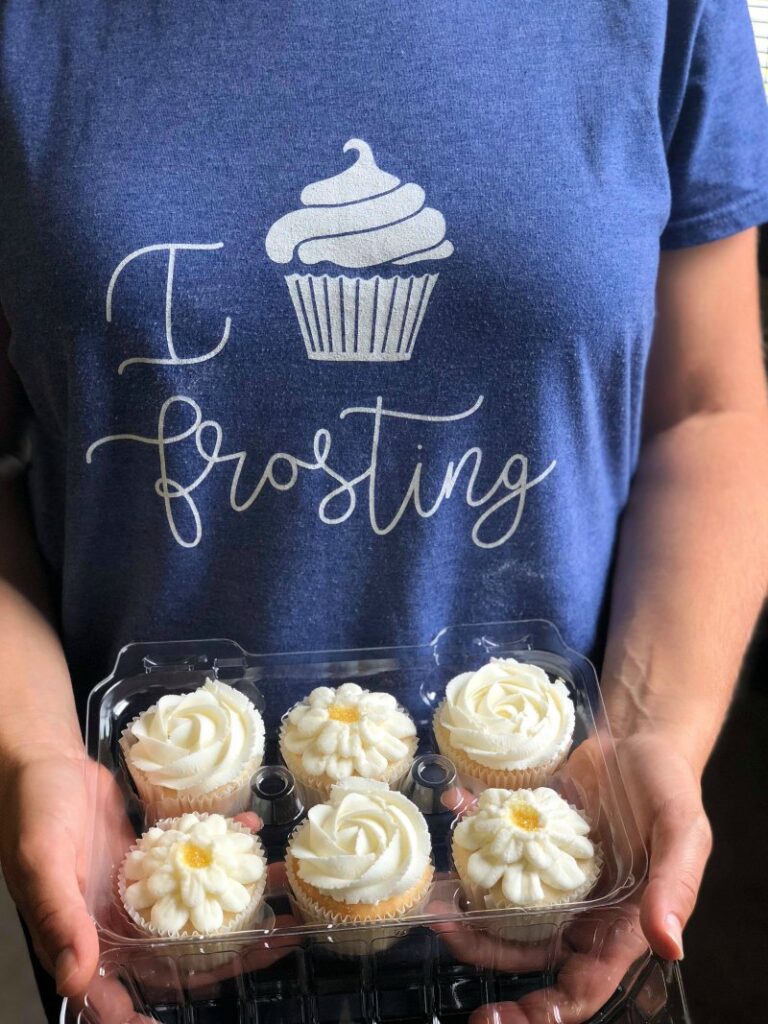 Hands holding container of cupcakes with buttercream flowers on top.