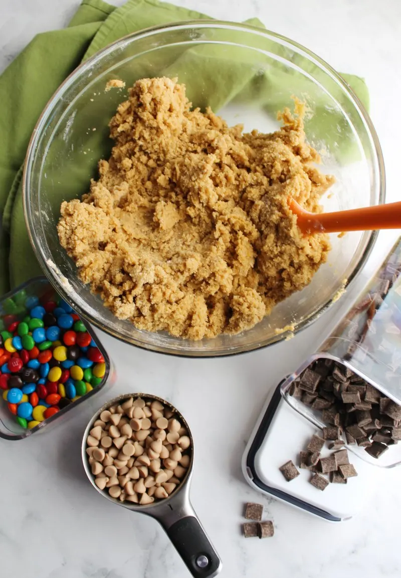 mixing bowl of blondie dough with container of chocolate chunks, m&ms and salted caramel chips in measuring cup.