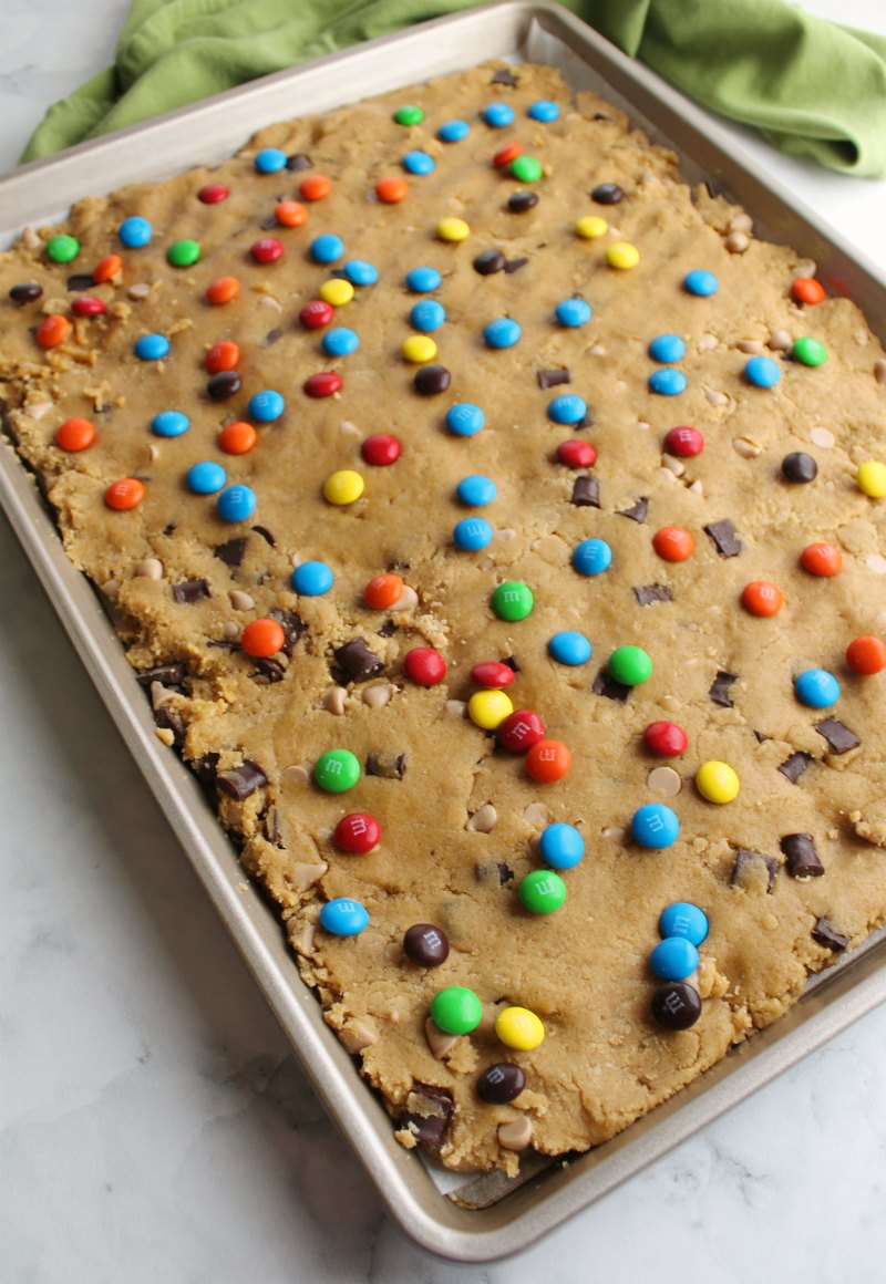 blondie dough loaded with salted caramel chips and chocolate chunks spread in half sheet pan with M&M candies scattered over the top.
