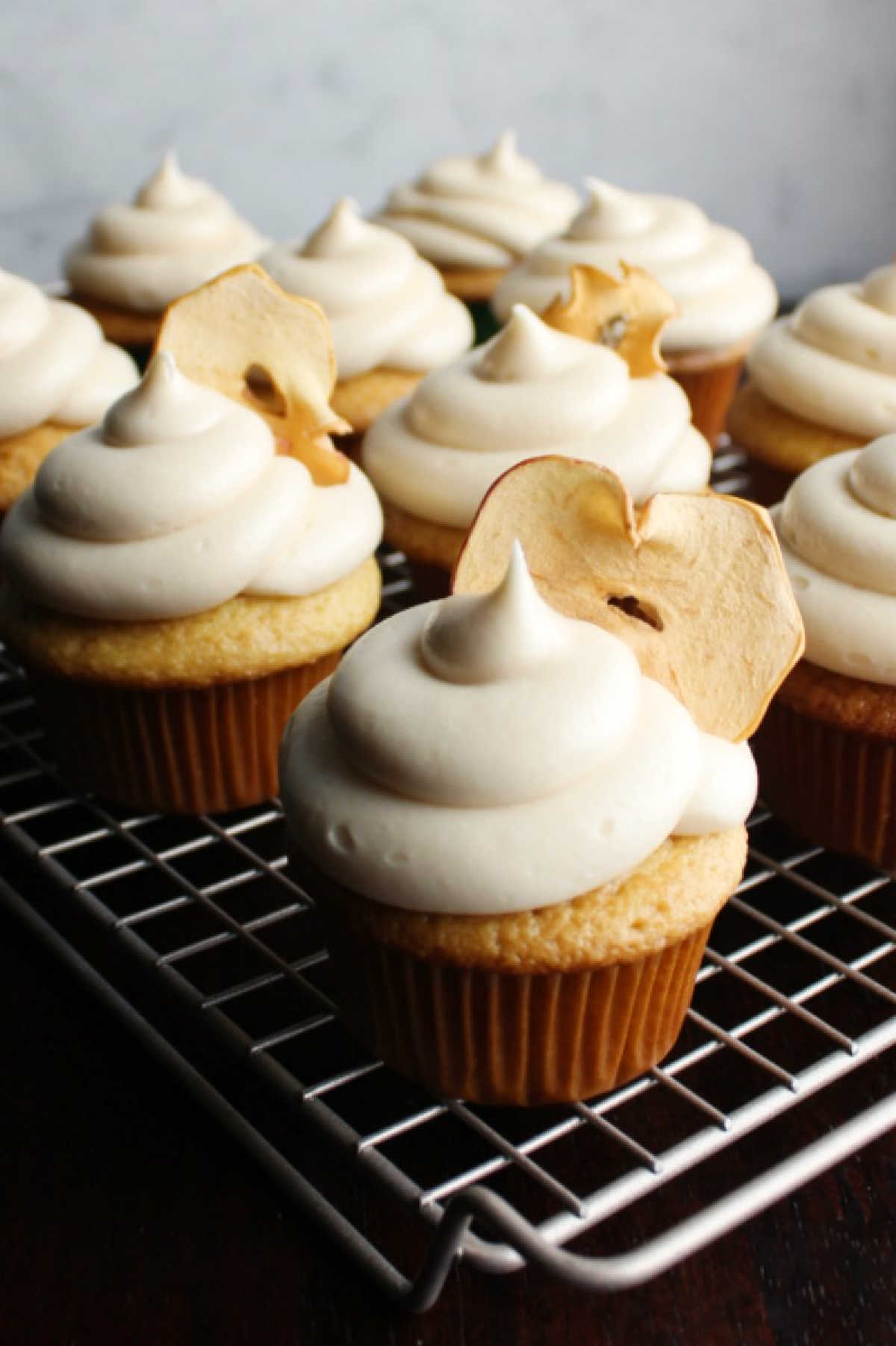 cooling rack loaded with apple cupcakes topped with caramel cream cheese frosting and apple chips.