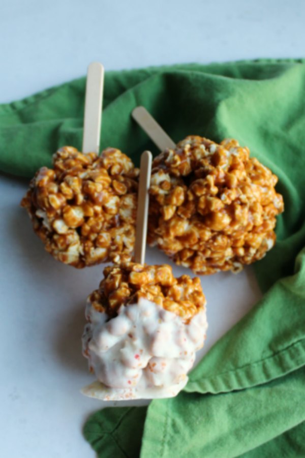 3 caramel apple popcorn balls on sticks with one dipped in white chocolate.