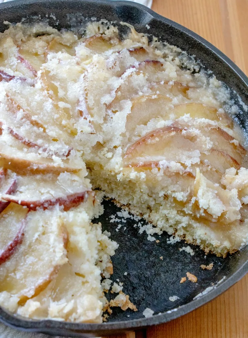 cast iron skillet full of dutch apple cake with buttery topping with one slice missing.