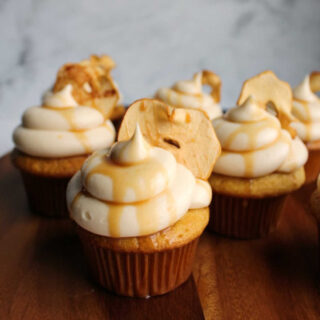 close up grouping of cozy apple cupcakes topped with caramel cream cheese frosting and apple chip and a drizzle of caramel sauce.
