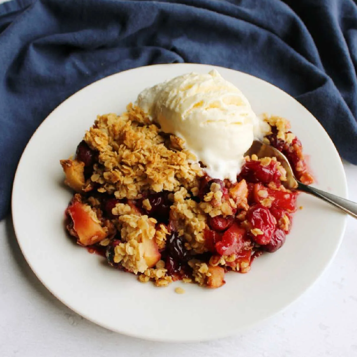 Dessert plate filled with cranberry apple crisp topped with golden oat topping and a melty scoop of vanilla ice cream.