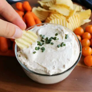 bowl of creamy cream cheese clam dip with chip being dipped in baby carrots and cherry tomatoes in the background.