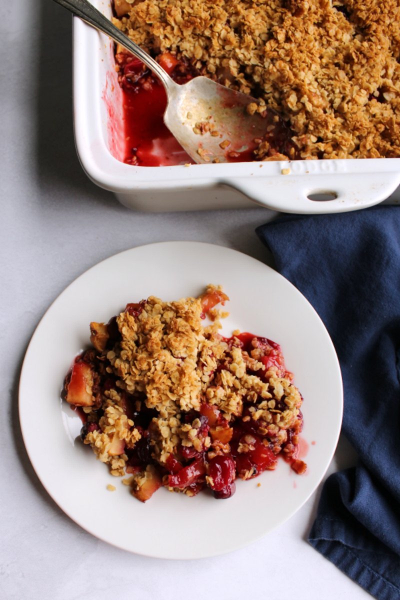 Small plate with serving of cranberry apple crisp with pan and antique berry spoon in the background.