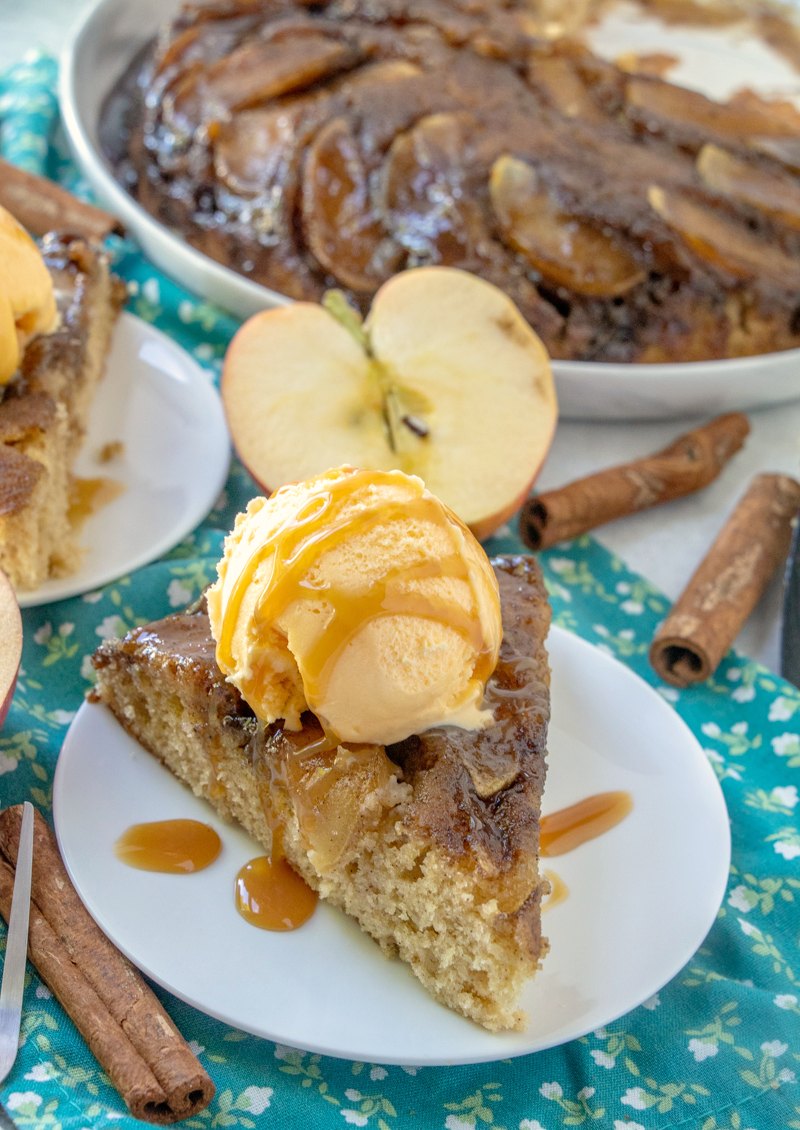 Slice of caramel apple upside down cake with cinnamon sticks and a cut apple nearby.