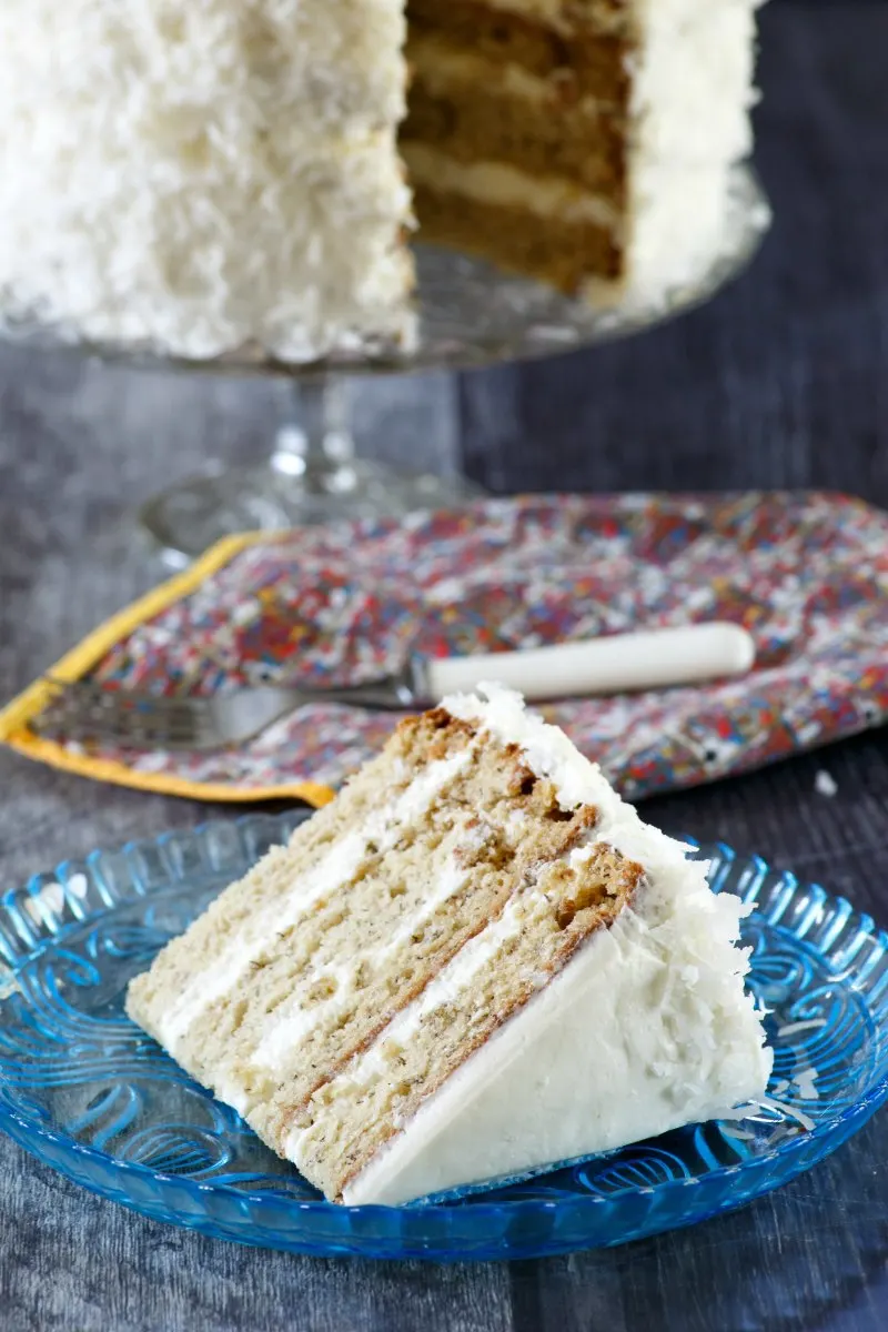 slice of layered banana cake with white frosting and coconut with remaining cake in background