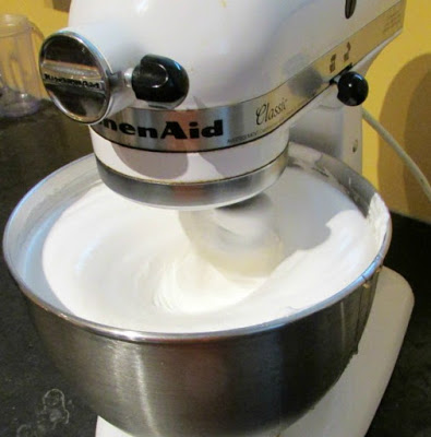 stand mixer running with bowl absolutely full of bright white fluffy frosting
