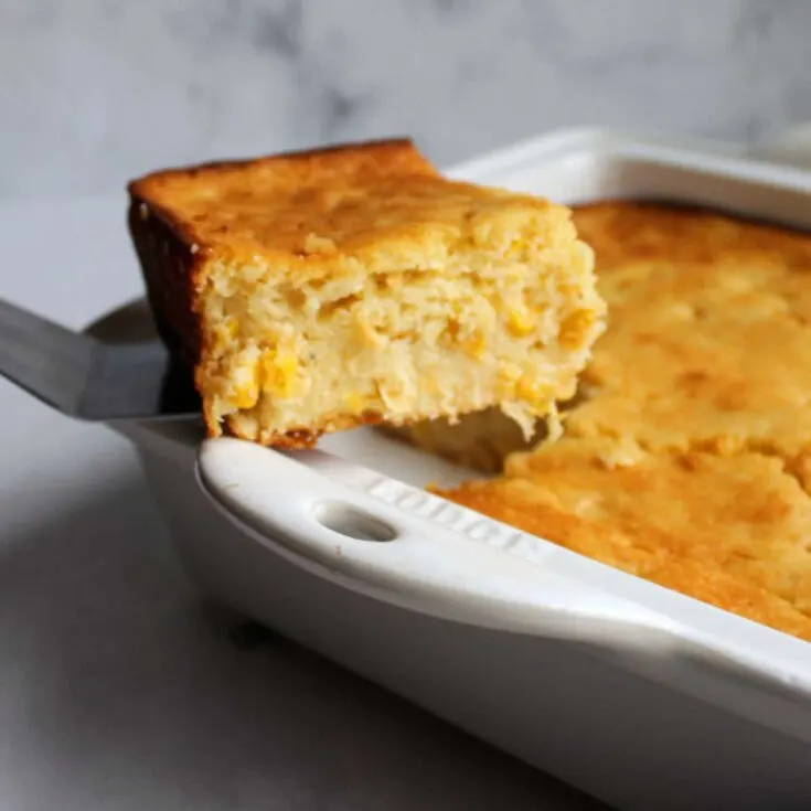 first slice of corn casserole being lifted out of casserole dish showing soft cornbread like center with dots of corn inside.